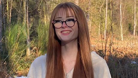 Brianna Ghey, with long red hair and black glasses, standing in a forest with trees around her