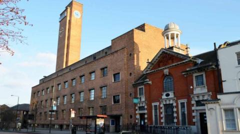 A 1930s Art Deco five storey red brick building with a tall clock tower. An older brick building topped with a cupola is to the right. 