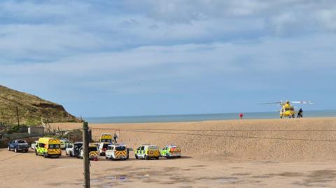 Six emergency vehicles, including an ambulance and the coastguard, are parked in a car park next to East Beach. The yellow air ambulance helicopter is sitting on the beach to the right of the photo. The sea is behind it and the cliffs are rising up on the left side of the image.