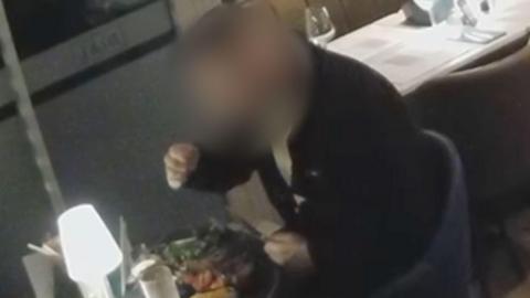 A still image from CCTV showing a man who appears to be pulling something from his hair. He is sitting in a restaurant and is wearing a black jacket.