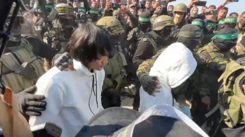 Two people in white hoodies are escorted by soldiers through a crowd