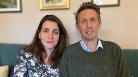 Andrew Davies (right) and his wife Milica. They are sitting on a green sofa and pictured from the chest up. She has long dark hair, is wearing a black and white marble-effect dress and drop pearl earrings. He has on a blue shirt with the collar unbuttoned and a dark green jumper over the top.
