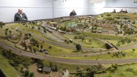 The railway has tracks, green fields, houses and there are people in the background.