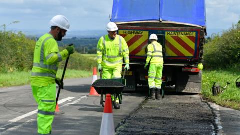 Three workmen in hard hats and wearing green, hi-vis jumpsuits are in front of a highway maintenance vehicle. One is holding a brush and another is pushing a wheel barrow full of tarmac. 