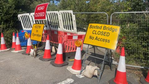 A road has been closed and cordoned off using metal and plastic fending. A row of orange traffic cones has been put in place to further block off access to the road. In front of these cones are metal signs. On the left there are two signs stacked on top of each other. At the bottom is a yellow metal sign with black writing which says, CCTV In Operation. On top of it, is a red metal sign with white writing which says, ROAD CLOSED. On the right of the picture there is a yellow metal sign which says, Boxted Bridge CLOSED No Access.