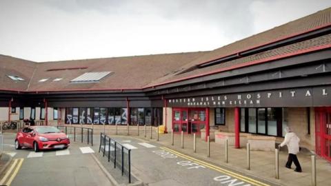 The entrance of the Western Isles Hospital in Stornoway with a car passing by and a pedestrian walking towards the front door