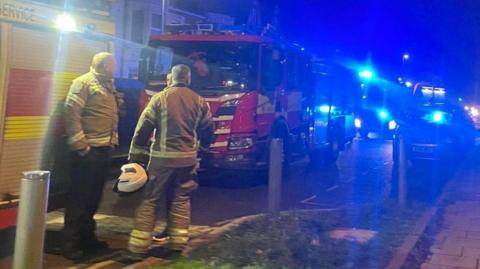 Two firefighters are standing in a road with their backs turned. At least three fire trucks are visible. It is dark.