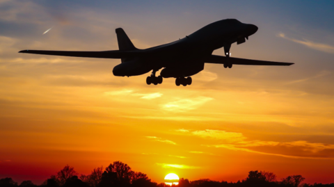The aeroplane is silhouetted in the sky against a stunning orangy-yellow sunset with the tops of a line of trees at the bottom of the picture and the sun disappearing on the horizon