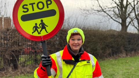 Maureen Seaton standing outside Coatham Primary School. She is wearing a bright hi-vis coat and hat. She has short red hair and is smiling at the camera. She is holding her lollipop stop sign.