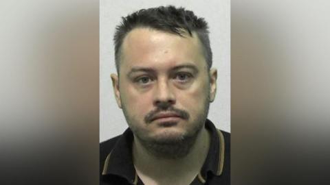 Mugshot of Aaron Keetley. He has scruffy short dark hair and a black moustache and beard,