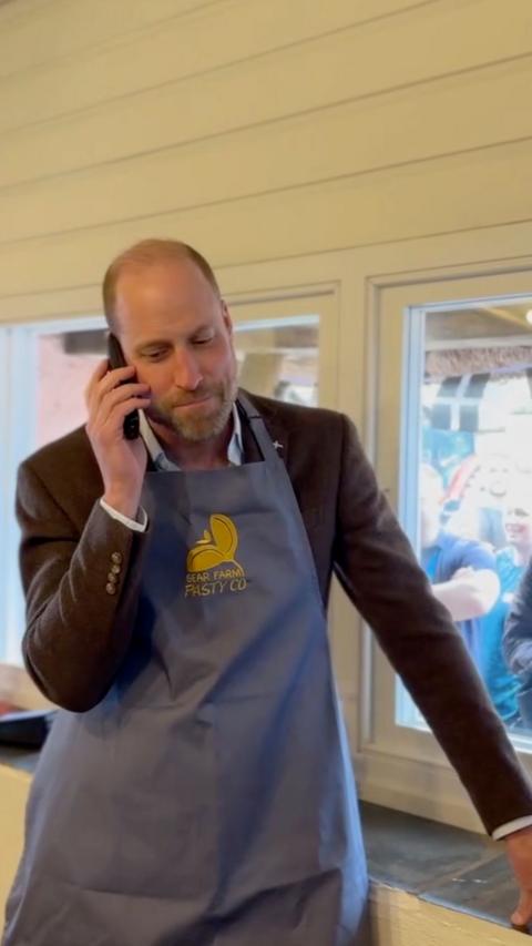 Prince William holding his phone up to his ear. He is wearing an apron and suit.
