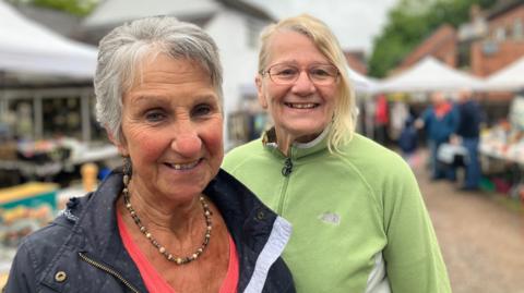 Two women look at the camera, smiling. One has short grey hair and a dark blue jacket, the other has longer hair and glasses, and is wearing a fleece.