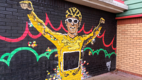 A colourful mural of Geraint Thomas wearing the yellow cycling jersey and helmet and white cycling glasses. Painted on a black background on a brick wall, with red and green swirls behind him.