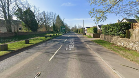 A road is pictured with no cars on it, and pavement and grassy verges either side.