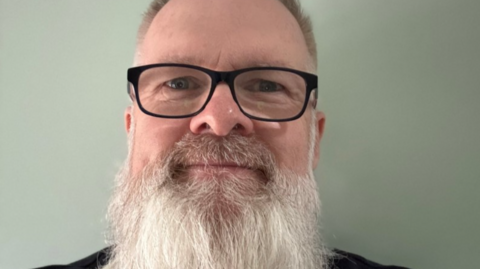 Chris has a long, white beard and is wearing glasses and smiling at the camera in a close-up selfie.