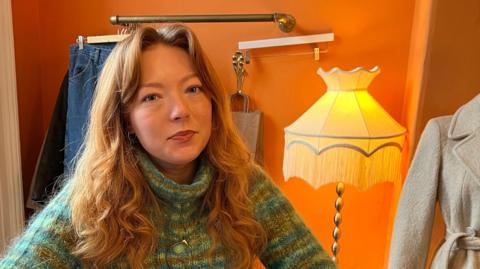 Laura Jackson. She is standing behind the counter of her shop and has long red hair and is wearing a green knitted jumper. A stylish coat and an ornate lamp are displayed behind her.