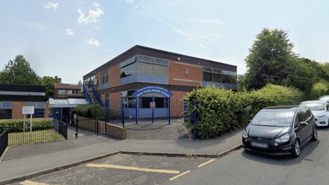 The entrance to Aston Fields Middle School is pictured from a street view. The entrance has a blue arch way with the name of the school written in white. The school building is predominately on two levels and made from red brick with blue windows.