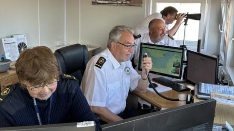 Three men wearing white shirts with black and yellow lapels are pictured looking out of the station windows. One is looking through a telescope while another is talking into a radio unit. A woman on the left is wearing a navy blue branded jumper with black and yellow lapels. She is looking at a computer screen in front of her.