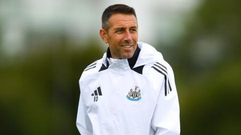 Ben Dawson smiles as he wears a white jacket with the Newcastle badge on the left and the Adidas logo on the right, with his initials underneath.