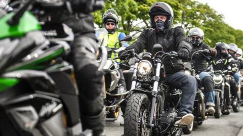Riders on motorcycles line up next to each other, they sit on their machines wearing leather jackets and helmets.
