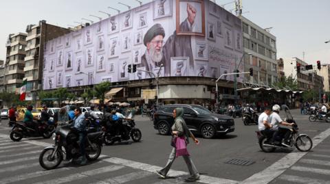 Iranians walk and drive in cars and on motorbikes near a huge poster of late Iranian supreme leader Ayatollah Ali Khamenei in a street in Tehran,