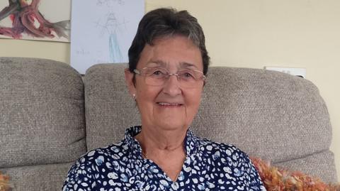 Woman with short black hair, wearing glasses and a navy and white patterned blouse. She is sitting on a cream sofa, an orange fluffy pillow can be seen over her left 