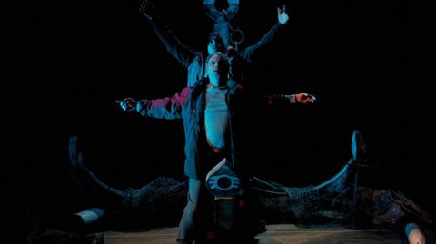 Two actors stand on stage dressed as fishermen, their arms outstretched, in front of a giant anchor.