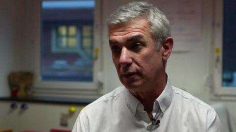 Hazlewood Primary School headteacher Stephane Bommel. He has short white hair and is wearing a white shirt, with a small microphone pinned to it. He is speaking to an interviewer out of shot and the background is blurred.