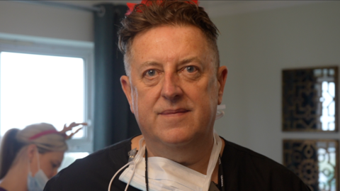 Dentist Stephen Denny with a PPE mask around his neck. He has black hair and is looking at the camera with a slight smile. He is standing inside a room at the care home and is wearing a black top.