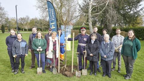 A group of councillors, Humber Forest workers and volunteers, and the lord lieutenant of East Yorkshire in ceremonial blue uniform stand with shovels in a park. The lord lieutenant is holding the tree to be planted.