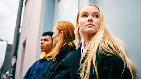Three young people lent against a building with a girl with long blonde hair looking away from her friends.