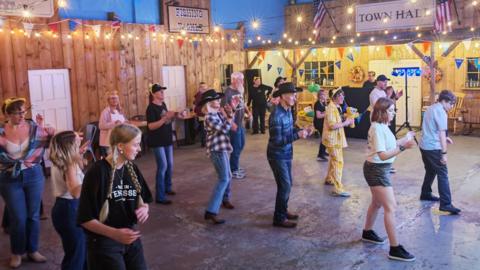 A group of line dancers in cowboy hats with the leader dressed in yellow