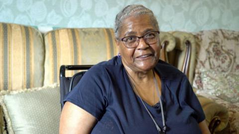 A woman with her hair in braids sits in front of her couch. She wears a navy t-shirt and smiles at the camera 