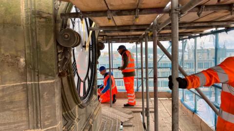 Three workers restoring a giant clocktower all wearing red high vis 