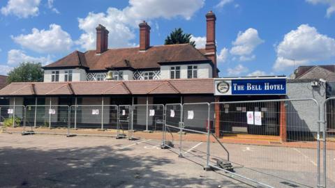 A white building with red roof with a sign stating The Bell Hotel in white lettering on a blue background above its doorway. The site is cordoned off by metal fencing
