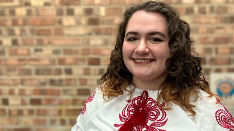 Yana Kozah is standing in front of a red brick wall. She is wearing a white shirt with red embroidery. Her hair is curly and brown. She is smiling at the camera.