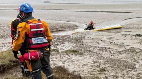 Coastguard members and Bay and Rescue members wearing brightly coloured diving suits have laid out a yellow stretcher by a sandy ditch on a beach to remove the two sheep.