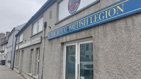 A picture showing the Royal British Legion building in Tandragee. It is a grey building with white doors, with a sign above it. The sign is blue with white writing that says 'The Royal British Legion'.
