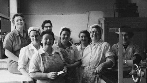 Black and white photograph of eight women posing to the camera in a factory room. They are all smiling. Some are wearing head coverings. Others are wearing aprons. Most are wearing jumpsuits. 