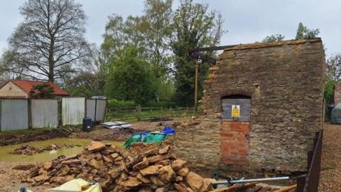 A old brick wall partly standing and partly in rubble. There is a hazard warning sign on the wall. There is water and soil on the floor and rubble bags. There is a fence, trees and brick building in the background.