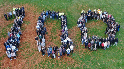 People stood on a lawn, making the shape of the number 100, seen from an aerial vantage point.