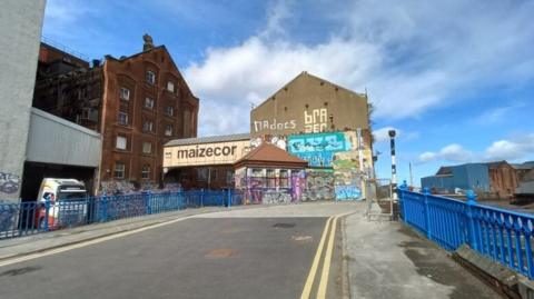 A warehouse to the left of the picture has a covered bridge coming out of it with "Maizecorn" printed on it.  The adjacent building has grafiti on it and the railings on the side of the road are painted bright blue.