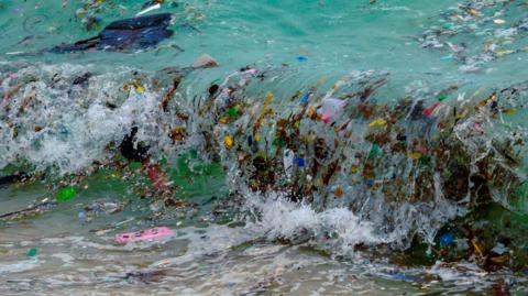 A wave crashes to shore carrying plastic pollution