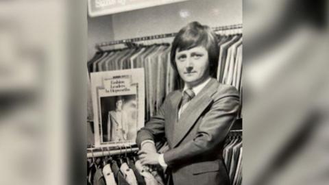 A black and white photo showing a man in a suit stood in a clothing shop surrounded by suit jackets. He is clean shaven and has a mod haircut style. 