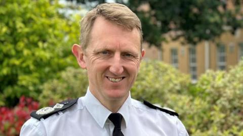 A man smiling at the camera wearing police uniform. He is wearing a white shirt with a black tie with police badges sewn on to black lapels on his shoulders. he is stood outside in front of greenery trees and bushes 