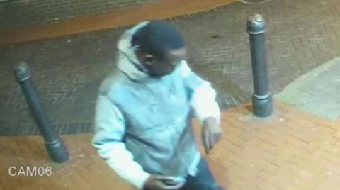 A man in a grey hoodie, apparently looking at his wrist, is standing on a street with bollards behind him.