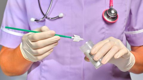 A nurse in a purple uniform places a sample, on the end of a green stick, into a clear and colourless tube. She has a stethoscope around her neck and wear white gloves.  