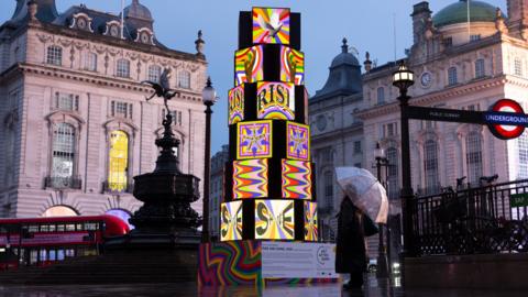 A brightly lit, multi-coloured cube sculpture stands in Piccadilly Circus at dusk, with a person holding an umbrella on wet pavement and historic buildings and a London Underground sign in the background.