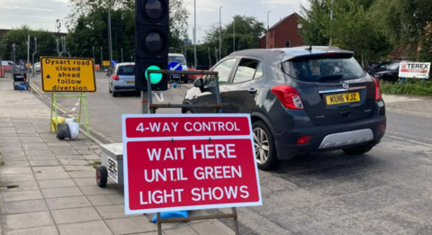 A red traffic sign says "4-way control - wait here until green light shows".  The light is on green and cars are weaving through past another yellow sign that says "Dysart road closed ahead follow diversion".