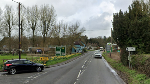 The A350, running through Longbridge Deverill near Warminster. There is grass either side of the carriageway and a junction to the left hand side.
